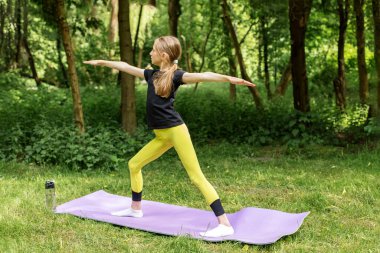 Atletik kıyafetli genç kız yeşil orman ortamında yoga minderinde savaşçı pozu veriyor. Denge ve farkındalığa odaklanıyor..