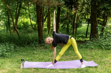 Genç bir kız, parkta mor bir paspasın üzerinde yoga pozu veriyor. Çevresi yeşil ağaçlarla çevriliyken, esnekliği ve dengesi üzerinde çalışırken temiz havanın ve doğal ışığın tadını çıkarıyor..