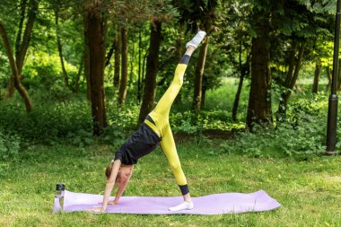 Genç kız, bir bacağı ormanda yüksekte yükselmiş, güç, esneklik ve kontrol odaklı zor bir yoga pozisyonu uyguluyor..