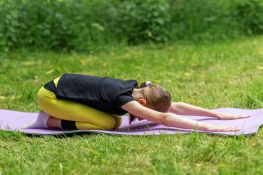Serin bir açık hava ortamında, bir çocuk mor bir minderin üzerinde yoga pozu veriyor, konsantrasyon ve dengeyi gösteriyor. Çimenli alan bu fitness aktivitesi için huzurlu bir zemin sağlıyor..