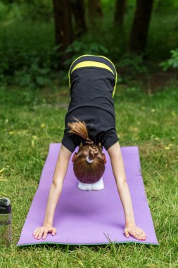 Küçük bir çocuk, ağaçlarla çevrili bir parkta mor bir paspasın üzerinde aşağıya doğru köpek yogası yapıyor. Hava güneşli görünüyor, pratik için neşeli ve huzurlu bir atmosfer yaratıyor..