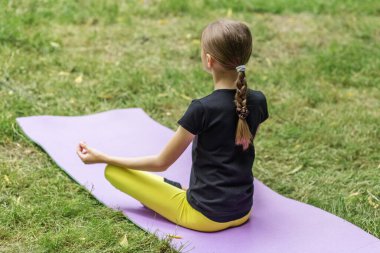 Bir çocuk, dışarıda huzurlu bir yerde minder üzerinde yoga yapıyor. Meditasyon yaparken denge ve konsantrasyon üzerine yoğunlaşır, rutin hareketleri sırasında zindelik ve sağlığını destekler..