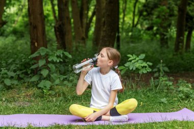 Genç kız parkta yoga minderinde oturur, su şişesi içer, dışarıda spor yaptıktan sonra dinlenir ya da egzersiz yapar..
