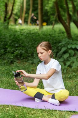 Genç kız parkta yoga minderinde oturur, su şişesini açarken, açık hava fitness ve egzersiz seanslarından sonra dinlenir..