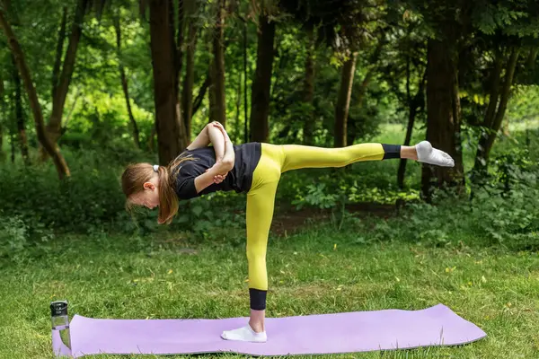 Ergen kız, yemyeşil bir ormanda mor bir paspasın üzerinde yoga pozu veriyor. Etrafı ağaçlarla çevrili, dikkatlice odaklanırken zarafetle dengede duruyor, dışarıda huzurlu bir ortamın tadını çıkarıyor..