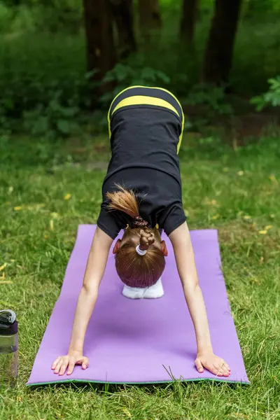 Küçük bir çocuk, ağaçlarla çevrili bir parkta mor bir paspasın üzerinde aşağıya doğru köpek yogası yapıyor. Hava güneşli görünüyor, pratik için neşeli ve huzurlu bir atmosfer yaratıyor..