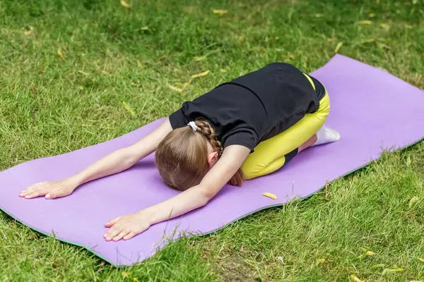 Bir çocuk, çimenli bir alanda mor bir minder üzerinde yoga pratiği yapıyor. Genç yogi, doğada esneme hareketleri yaparken denge ve konsantrasyonu korumaya odaklanır..