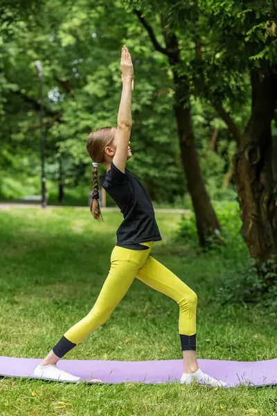 Bir çocuk parkta yoga pozu veriyor denge ve odaklanmayı gösteriyor. Genç pratisyen, yeşillikle çevrili mor bir paspasın üzerinde uzanıyor, fitness ve refahı teşvik ediyor..