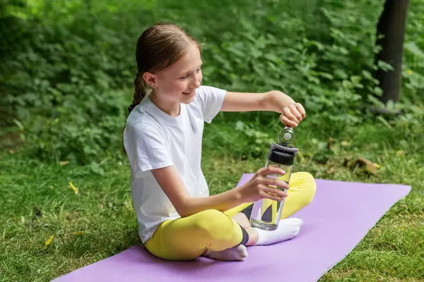 Genç kız parkta yoga minderinde oturur, su şişesini açarken gülümser, açık hava fitnesinden sonra dinlenir ya da egzersiz yapar..