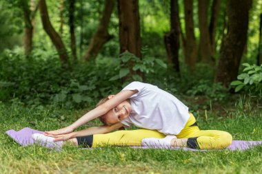 Genç bir kız, ormanın minderinde oturup yoga yapıyor. Gülümsüyor ve dışarıda huzurlu bir şekilde egzersiz yapıyor..