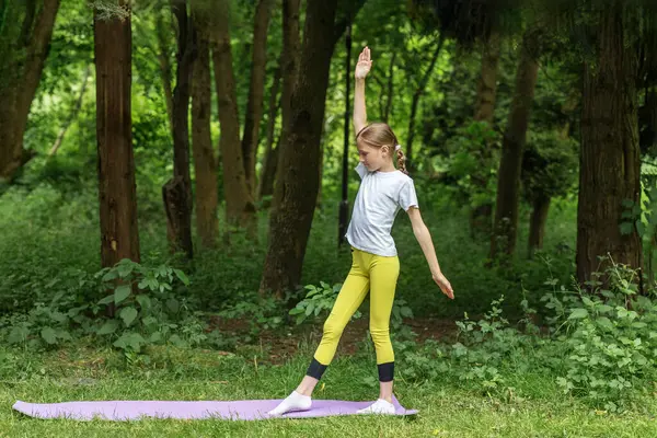 Bir ergen, yeşil ağaçlardan oluşan bir gölgeliğin altında yoga pozu verir. Çocuk bu açık hava egzersizi sırasında esneklik ve dengeyi artırırken dikkatle odaklanır..