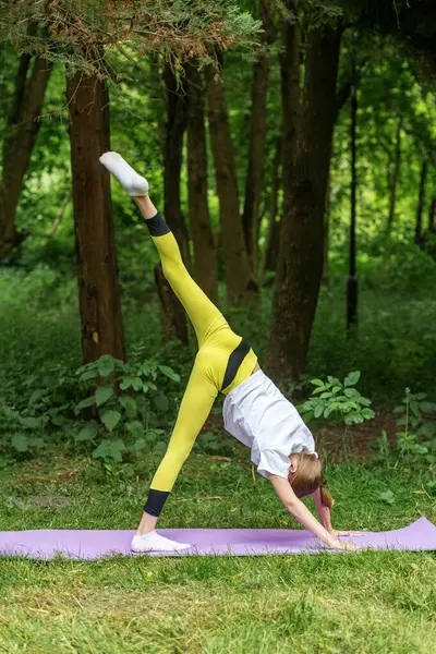 Genç kız, bir bacağı ormanda yüksekte yükselmiş, güç, esneklik ve kontrol odaklı zor bir yoga pozisyonu uyguluyor..