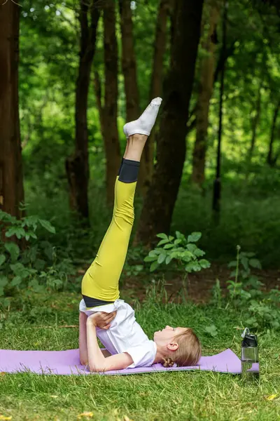 Bir ergen, ağaçlarla çevrili yemyeşil bir alanda yoga pozu veriyor. Denge ve konsantrasyona odaklanan çocuk zindelik ve refah için esneme hareketleri yapıyor..