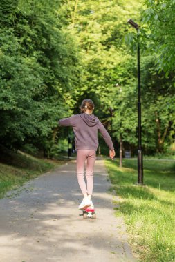 Genç kız, güneşli bir günde ağaçlarla kaplı sakin bir park yolunda kaykay sürerken denge kuruyor..