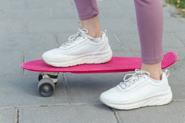 Açık hava ortamında, asfalt kaldırımda parlak pembe bir kaykay süren beyaz spor ayakkabı giyen birinin yakın çekimi..