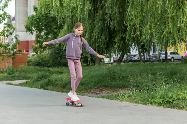 Mor aktivite kıyafetli bir genç kız pembe bir kaykayla yeşil ve park etmiş arabaların yanındaki kaldırımda güven içinde geziyor..
