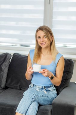 Keyifli genç bir kadın, rahat bir ev ortamında kanepede oturuyor, elinde bir kahve fincanı, mutlu ve rahat görünüyor..