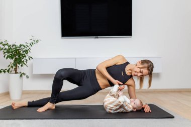 Anne, bebeğiyle meşgul olurken yoga minderinin üzerinde bir yan tahta pozu tutuyor. Egzersiz yapıyor ve evde şakacı bir bağ kuruyor..