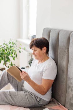 Yaşlı bir kadın rahat, aydınlık bir ev ortamında ev bitkileriyle yatağında oturup sessizce kitap okumaktan zevk alıyor..