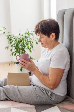 Yaşlı bir kadın yatakta oturuyor, sıcak bir içki tutuyor ve parlak, rahat bir yatak odasında açık bir kitapla rahatlıyor..