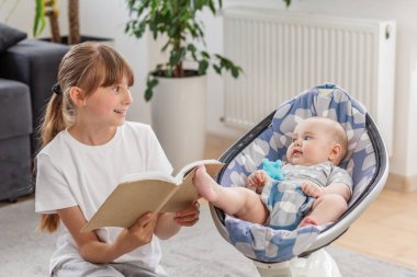Mutlu genç kız, sallanan erkek kardeşine yüksek sesle kitap okur, sıcak bir yuva ortamında bağlanma anını paylaşır..
