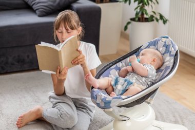 Genç kız, bebek kardeşinin yanında otururken sesli bir şekilde kitap okuyor. Modern bir bebek rockçıda, rahat bir oturma odasında..