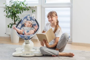 Gülümseyen genç kız, güneşli bir odada sallanan kardeşinin yanında otururken bir kitap okur..