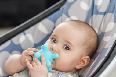 Sevimli bebek sallanan bir kayanın içinde uzanır, parlak mavi bir diş çıkaran oyuncağı tutar ve çiğnerken büyük meraklı gözleriyle bakar..