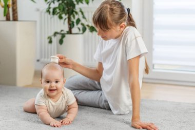 Mutlu bebek halının üzerinde yatarken ablamız nazikçe başını ovalıyor, hassas ve neşeli bir aile bağı kuruyor..