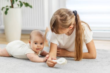 Genç bir kız ve bebek yerde uzanıp, yumuşak bir bebek fırçasıyla etkileşime girerken birlikte sakin ve eğlenceli bir anı paylaşıyorlar..