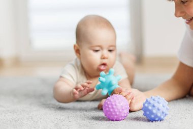 Bebek göbeğinde, bir bakıcı tarafından tutulan parlak mavi duyusal topa uzanıyor, halının üzerinde renkli desenli oyuncaklarla çevrili..