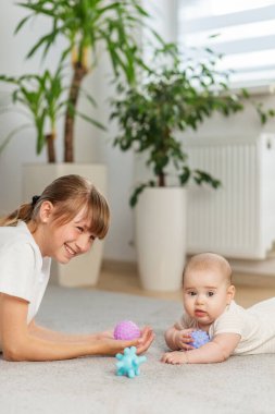 Gülümseyen kız, renkli duyusal oyuncaklar kullanarak halının üzerinde uzanan bebekle meşgul oluyor, rahat bir evde neşeli bir bağ kurma anı yaratıyor..