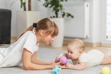 Genç kız, halı döşeli bir zemin üzerinde oyun saatinde dokulu mor bir topu incelerken erkek kardeşinin ellerine nazikçe rehberlik ediyor..
