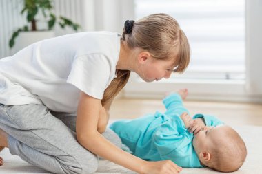 Genç kız kardeşinin yanında diz çöker, yere uzanır, gülümser ve parlak bir odada neşeli bir etkileşim içinde olur..