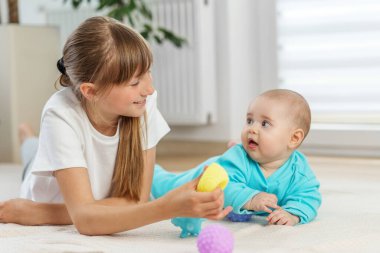 Küçük kız, sarı desenli bir oyuncak tutarken kardeşine gülümsüyor. İkisi de parlak bir odada, rahat bir halının üzerinde yatıyor..