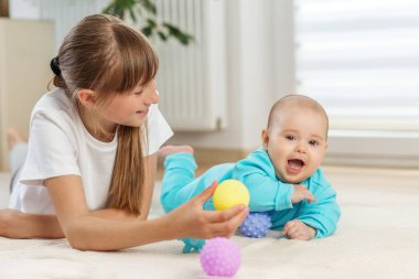 Bebek turkuaz kıyafetli, gülümseyen ve gülen bir kız kardeş yumuşak bir iç halının üzerinde renkli duyusal toplarla oynarken...