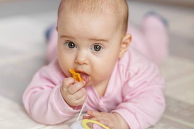 Meraklı bebek pembe giysiler içinde yerde yatar, oyuncağını yalarken aynalı ve şekilli yumuşak eğitici bir oyuncağı keşfeder..