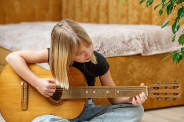Sarışın kız, rahat bir yatak odası ortamında yerde oturmuş, parmak yerleştirme ve akorlara odaklanmış halde akustik gitar çalıyordu..