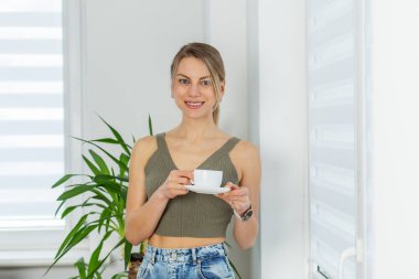 Yeşil bluzlu ve kotlu genç bir kadın sıcak bir gülümsemeyle beyaz bir kahve bardağını parlak bir pencerenin yanında tutarken..
