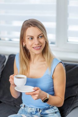 Sarışın kadın elinde beyaz bir kahve fincanı tutarken gülümsüyor ve yan tarafa bakıyor, evinde kanepede dinleniyor..