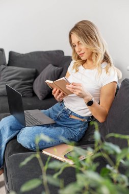 Dizüstü bilgisayarı ve defteri olan genç bir kadın rahat bir ortamda dijital iş ve el yazısıyla yazılmış notlar arasında gidip geliyor..