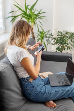 Ev bitkileriyle çevrili, iş ve rahatlamayı birleştiren parlak bir evde akıllı telefon ve dizüstü bilgisayar kullanan sıradan bir kadın..