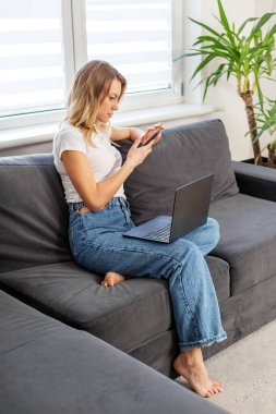 Kanepede çıplak ayakla oturan genç bir kadın akıllı telefon ve dizüstü bilgisayar kullanıyor. Parlak bir ev ortamında doğal ışık ve bitkilerle çalışıyor..