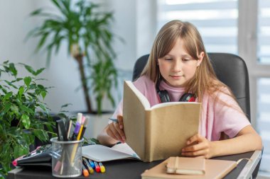 Kulaklıklı genç bir kız, temiz bir masada, iç mekan bitkileriyle çevrili kalemler ve defterlerle dikkatle kitap okur..