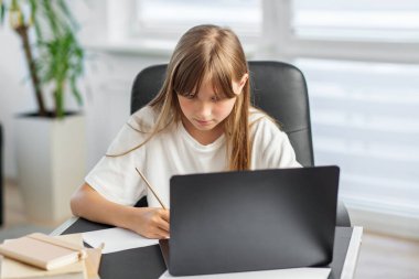 Beyaz gömlekli ciddi bir kız, online çalışmalar sırasında dizüstü bilgisayara bakarken masa başında notlar yazıyor..