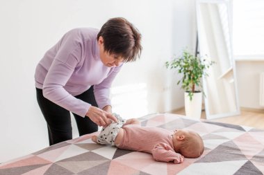 Parlak, modern bir yatak odasında, bebek pantolonu giymiş yaşlı bir kadın, sevgi ve günlük bakım gösteriyor..