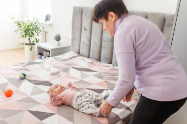 Yaşlı bir kadın, bebeği nazikçe yatağa yatırıyor. Parlak, rahat yatak odasında oyuncaklarla çevrili, sevgi ve şefkat gösteriyor..