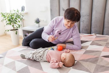 Yaşlı bir kadın yatağa uzanıp bebekle oynuyor. Parlak odada yumuşak oyuncaklar kullanıyor. Sevgi, etkileşim ve bağ gösteriyor..