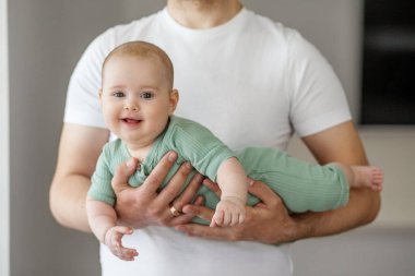 Evde baba tarafından nazikçe kucaklanırken yeşil elbiseli neşeli bir bebek gülümsüyor. Doğal ışıkla aydınlık ve neşeli bir aile anı..