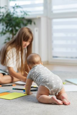 Puantiyeli bebek, renkli kağıtlar ve işaretlerle çevrili, çizim yapan ablasına doğru sürünüyor..